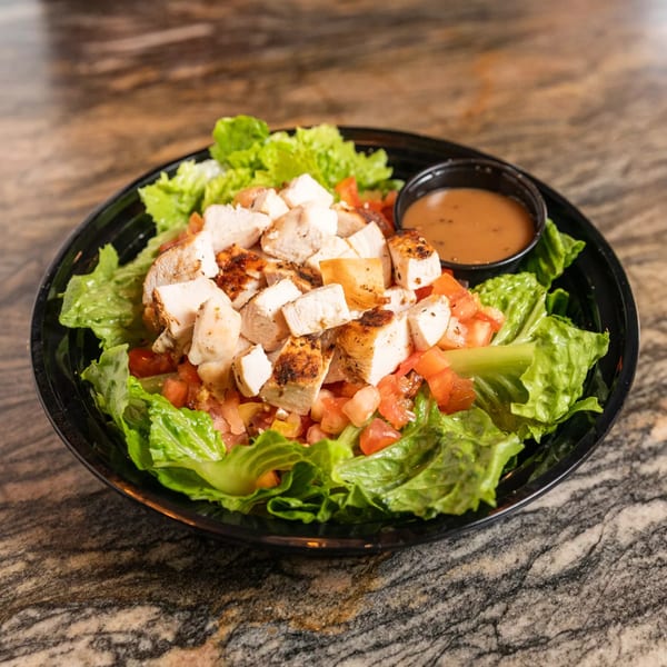 A grilled chicken salad with lettuce, diced tomatoes, and a side of dressing in a black bowl.