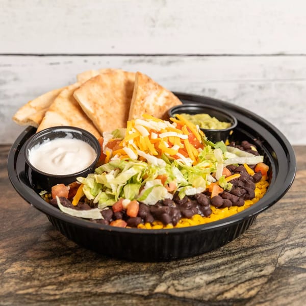 A black bowl of rice, black beans, lettuce, cheese, and tomatoes, with sides of sour cream, guacamole, and pita bread.